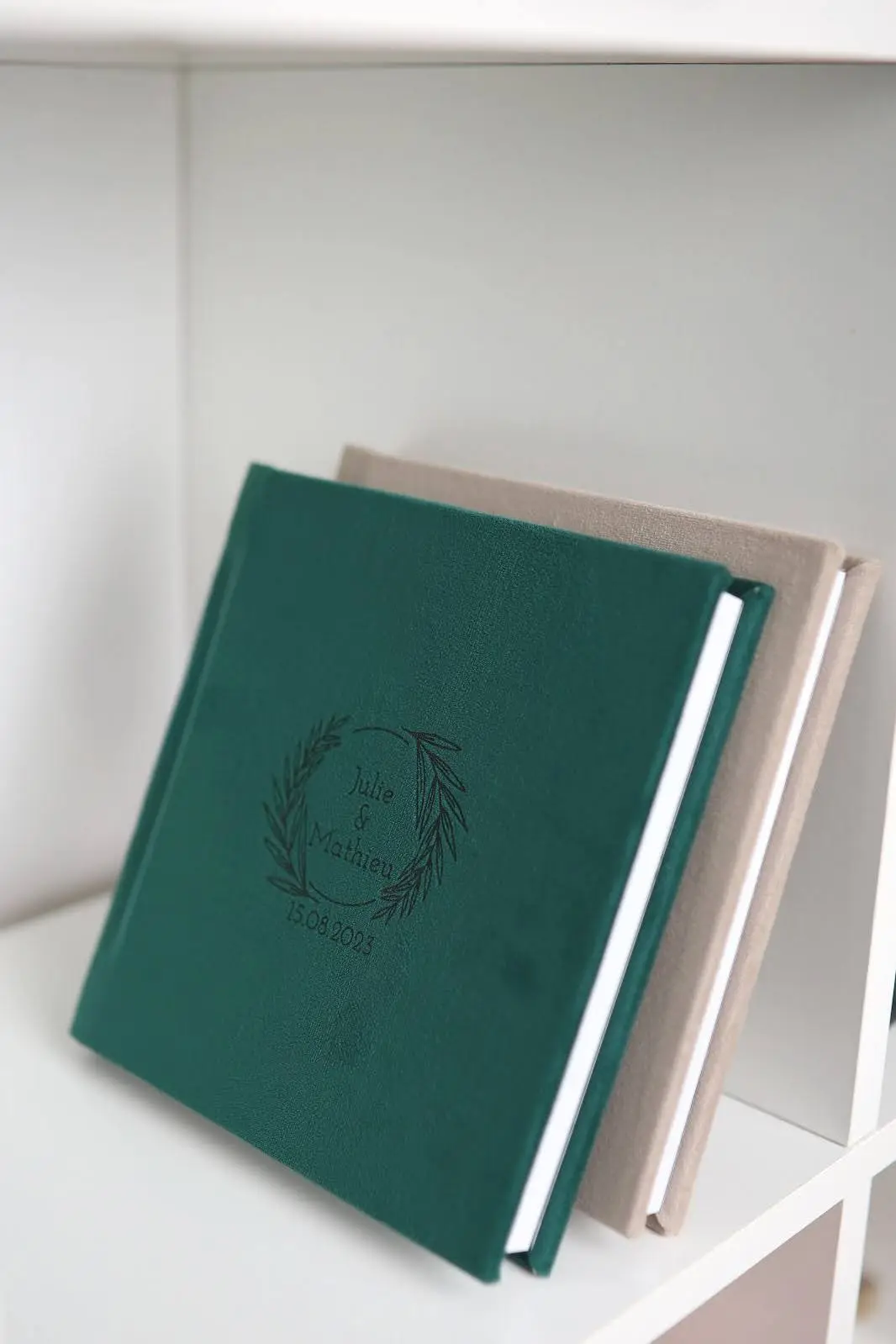 Two hardcover photo albums displayed on a white shelf, with the front one in dark green featuring “Julie & Mathieu 15.08.2023” embossed in a wreath design. The second album behind it is beige, both elegant and neatly arranged.