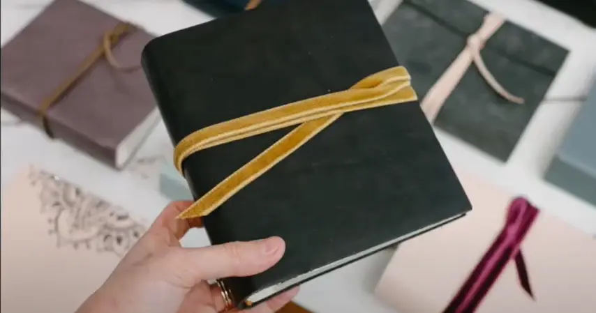 Hand holding a black brag book photo album wrapped with a yellow velvet ribbon, surrounded by other ribbon-tied albums.
